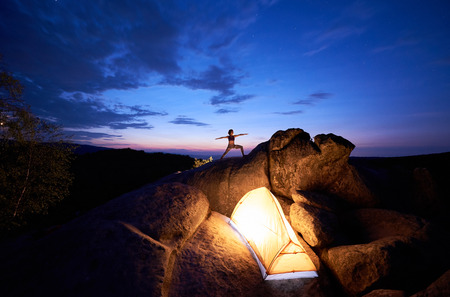 Camping on rock formation. Brightly lit tourist tent and silhouette of slim tourist girl doing yoga exercises on mountain top against dark blue sky at sunset. Sport, tourism concept. Virabhadrasanaの写真素材