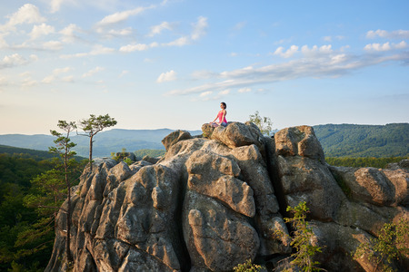 Athletic barefooted woman sitting in lotus pose on lit by summer sun top of huge rock on green mountains and clear blue sky background. Yoga, fitness and healthy lifestyle concept. Siddhasanaの写真素材