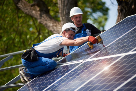 Professional technicians working with electrical screwdriver installing shiny solar photo voltaic panel to metal platform system on green tree thick foliage background.の写真素材