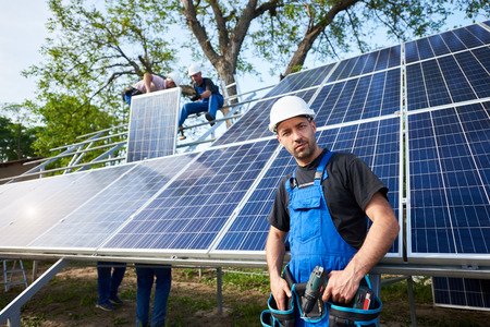Portrait of young confident engineer technician with electrical screwdriver standing in front of unfinished high exterior solar panel photo voltaic system with team of workers on high platform.の写真素材