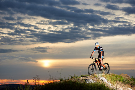 Professional cyclist riding mountain bike on rock hill with enthusiasm. Energetic and athletic man in sportswear, helmet and sport glasses. Beautiful background of evening sky and sun.の写真素材