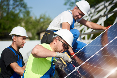 Three professional technicians installing solar photo voltaic panel to metal platform on blue sky background. Stand-alone solar system installation, efficiency and professionalism concept.の写真素材