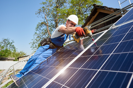 Technician installing solar photo voltaic panel to metal platform using screwdriver on bright blue sky background. Stand-alone solar panel system installation concept.の写真素材