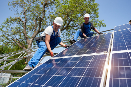 Two young technicians installing heavy solar photo voltaic panel on tall steel platform on green tree background. Exterior solar panel voltaic system installation, dangerous job concept.の写真素材
