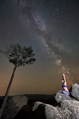 Beautiful summer night in mountains. Woman sitting alone on top of huge boulder doing yoga near pine tree, under clear sky with blinking stars and Milky way. Tourism and hiking concept. Siddhasanaの写真素材