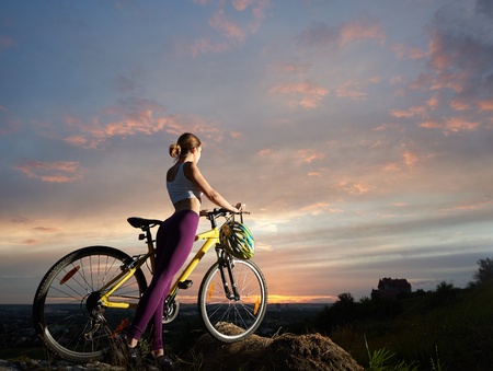 Slender woman with a mountain bike is standing on a hill under a beautiful sky at sunset against the background of the town in the distance. Helmet hangs on the handle of a bicycleの写真素材