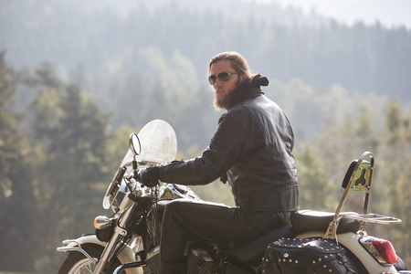 Back view of bearded motorcyclist with long hair in black leather jacket sitting on cruiser motorcycle on blurred background of spruce tall trees and hills on horizon. Active lifestyle and traveling.の写真素材