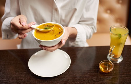 Crop of young female eating tasty orange cream soup. Woman in blouse enjoying tasty first dish in luxury restaurant. Client eating exotic meal on lunch. Concept of oriental food.の写真素材