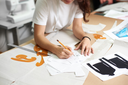 Young smiling female designer drawing pencil sketches of clothing sitting at big table with flat paper patterns, measuring ruler and curves on blurred background of tailor design studio interior.の写真素材