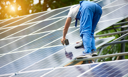 Construction worker connecting photo voltaic panel to solar system using screwdriver on shiny surface and lit by sun green tree background. Alternative energy and financial investment concept.の写真素材