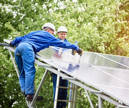 Team of two constructors connecting photo voltaic panel to stand-alone solar system platform using screwdriver. Alternative energy, professionally done job and profitable financial investment concept.の写真素材