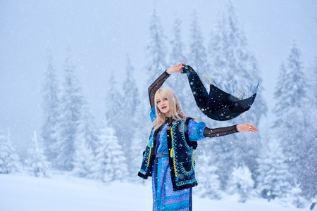 Attractive fashionable blond girl in long blue dress, embroidered warm sleeveless fur coat outdoor posing in deep clean snow on bright sunny winter day on blurred spruce trees copy space background.の写真素材
