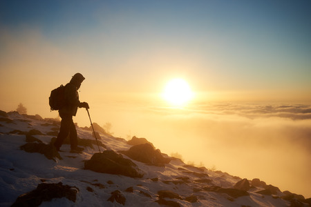 Tourist climber with backpack and trekking poles on rocky snowy mountain steep slope on copy space background of foggy valley filled with white puffy clouds, raising sun and bright blue sky at dawn.の写真素材