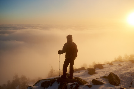 Back view of silhouette tourist hiker with backpack and hiking sticks on rocky mountain peak on copy space background of beautiful foggy valley filled with white puffy clouds at dawn.の写真素材