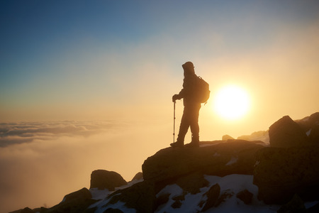 Silhouette of tourist hiker with backpack and hiking sticks resting on the top of rocky snowy mountain on copy space background of bright white raising sun and misty orange blue sky at dawn.の写真素材