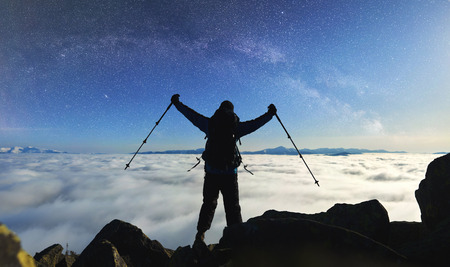 Back view of tourist hiker in winner posture on the top of mountain with trekking poles in raised arms on bright blue starry sky with Milky way at night, foggy valley filled with white clouds.の写真素材