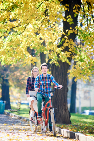 Young happy treveller couple in casual clothing, bearded man and blond woman cycling on modern tandem bicycle on lit by bright sun empty pavement in beautiful autumn park under tall trees.の写真素材