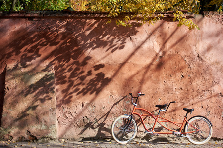 Modern comfortable orange tandem double bicycle with white wheels on paved empty sidewalk leaned against old plastered cracked red wall on bright sunny autumn day. Transport and traveling concept.の写真素材