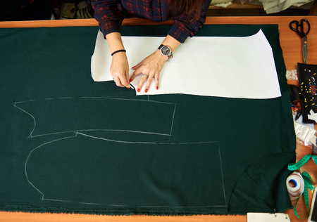 Tailor worktable in the studio. Close-up of woman hands making chalk markings of paper patterns on green fabric copy space surface before cutting details for future jacket or costume.の写真素材