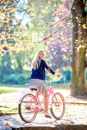 Young happy attractive blond long-haired woman in glasses, skirt and blouse cycling pink lady bike along sunny paved park alley on blurred golden yellow green bokeh foliage background.の写真素材