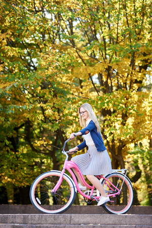 Side view of attractive blond long-haired woman in glasses, skirt and blouse cycling modern pink lady bike on lit by autumn sun park stairs on bright colorful golden bokeh trees background.の写真素材