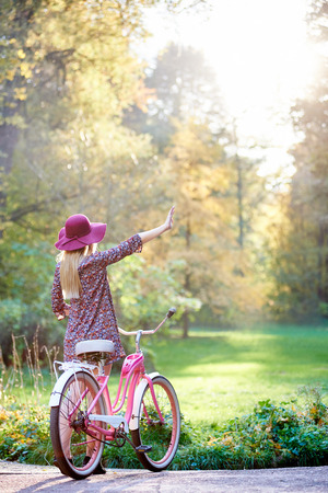 Back view of slim blond fashionable long-haired attractive girl in short dress and pink hat with raised arm at lady bicycle on paved summer park alley on beautiful green and golden trees background.の写真素材