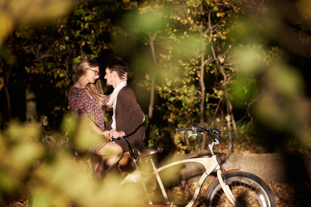 Happy romantic couple, bearded man and attractive girl close together at tandem double bicycle outdoors in autumn park on sunny clearing in the evening on blurred golden green bokeh background.の写真素材
