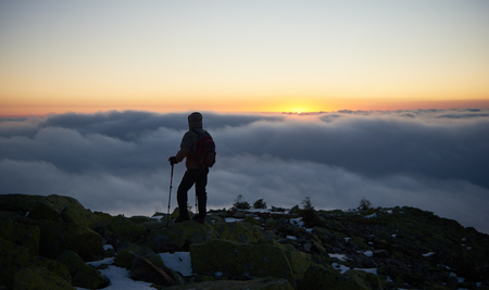 Back view of tourist in warm jacket with backpack and hiking sticks on rocky mountain peak enjoying view of valley covered with white clouds stretching to horizon and bright morning sky at sunrise.の写真素材