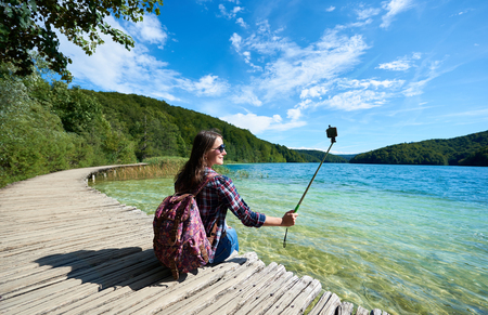 Colorful spring panorama of lit by bright sun green forest and clear blue lake. Young tourist woman in sunglasses and with backpack and camera sitting on wooden bridge enjoying beautiful view.の写真素材