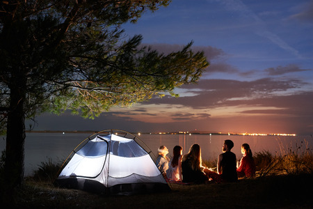 Back view of five tourists, women and man sitting on sea shore around bonfire at tent under tree on smooth water surface and blue evening sky at sunset background. Tourism and camping concept.の写真素材