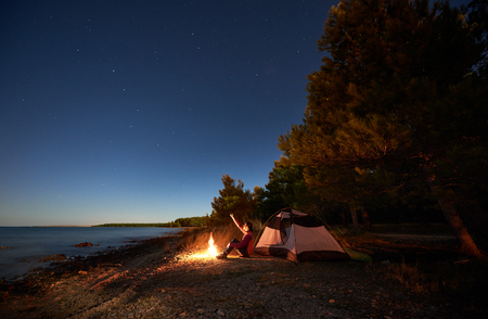 Night camping at sea shore. Woman hiker sitting in front of tourist tent at campfire, pointing to starry sky, enjoying beautiful view of clear blue water. Tourism and active lifestyle conceptの写真素材
