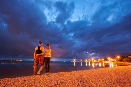 Back view of young couple, man and long-haired woman standing embraced on resort town pebble beach at dusk on calm blue water with reflected street lamps light background. Tourism and vacations.の写真素材