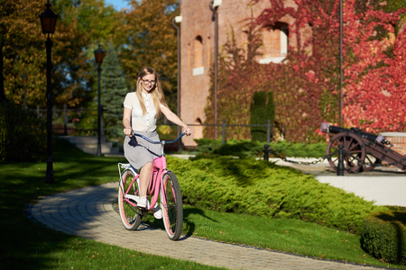 Active blond long-haired attractive tourist woman in glasses and casual clothing cycling modern pink lady bike on bokeh background of beautiful different green trees in the fall.の写真素材