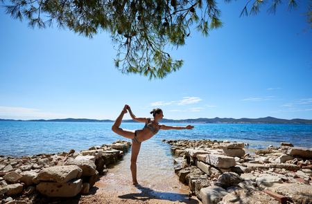 Young attractive suntanned woman in swimming suite making yoga pose near sea water by stony path under bright blue sky. Active lifestyle, summer holidays, recreation and vacations concept.の写真素材