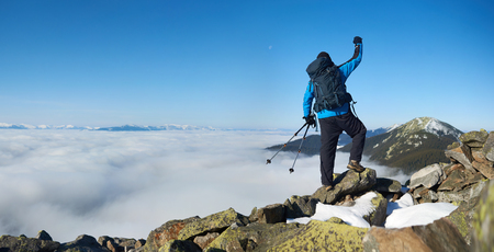 Back view of tourist hiker with backpack and trekking poles standing with raised arm on huge rocks on background of foggy valley filled with white clouds, distant mountain tops and blue sky.の写真素材