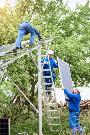 Installing of stand-alone solar photo voltaic panel system. Workers in hard-hats and blue overall lifting the solar module on metal platform. Alternative energy and professional construction concept.の写真素材