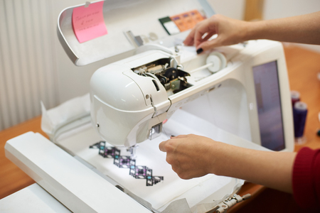 Close-up of woman hands filling thread in sewing machine on background of white textile detail with embroidered colorful geometrical pattern. Modern technology and fashionable clothing manufacturing.の写真素材