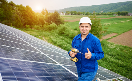Close-up of construction worker with screwdriver with thumb-up gesture on photo voltaic panel solar system shiny surface and lit by sun green fields background. Alternative energy concept.の写真素材