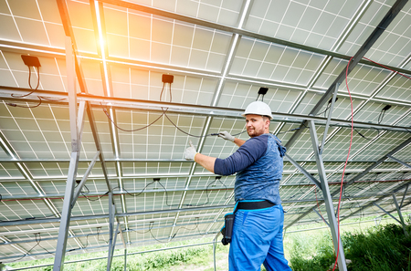 Young electrician in hard hat and rubber gloves wiring solar photo voltaic modules inside the new installed panel system on bright sunny day. Alternative energy and profitable investment concept.の写真素材