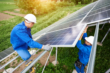 Installing of stand-alone solar photo voltaic panel system. Two technicians in hard-hats mounting big shiny solar module on platform on green summer view background. Alternative energy concept.の写真素材