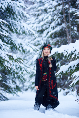 Attractive girl in dark blue embroidered long sleeveless sheepskin fur coat and kerchief with floral design outdoors in deep snow on sunny winter day on blurred background of snowy spruce trees.の写真素材