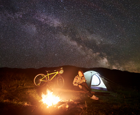 Young woman cyclist resting at night camping near burning campfire, illuminated tourist tent, mountain bicycle under incredible beautiful evening sky full of stars and Milky way. Tourism conceptの写真素材