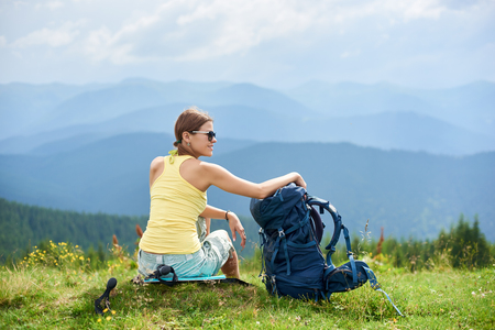 Back view of attractive woman tourist sitting on grassy hill with backpack, wearing sunglasses, enjoying summer cloudy day in the Carpathian mountains. Outdoor activity, tourism conceptの写真素材