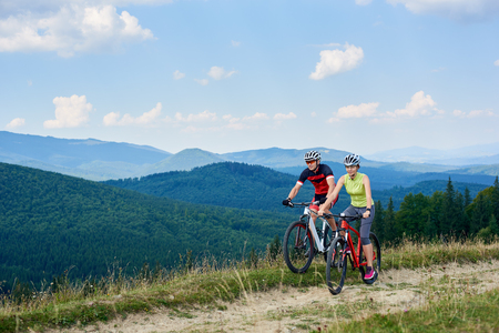Young bikers tourists, man and woman in professional sportswear riding bikes down grassy field road under beautiful bright blue sky on magnificent mountain range background. Active lifestyle concept.の写真素材
