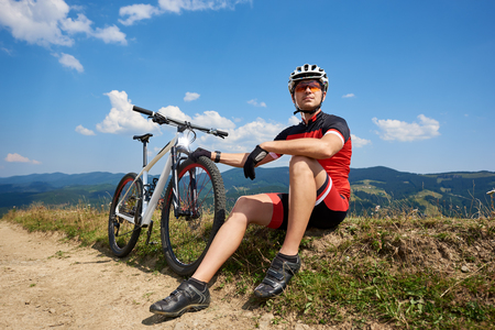 Young muscular sportsman biker in professional sportswear sitting near his bike on dusty roadside resting on bright beautiful mountains summer background. Active lifestyle and extreme sport concept.の写真素材