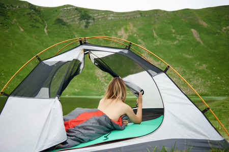 Back view of attractive naked female tourist lying in tent in sleeping bag, enjoying beautiful view of lake in the mountains. Camping lifestyle concept adventure summer vacations outdoorの写真素材
