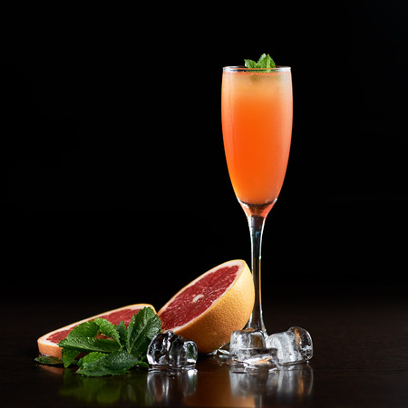 Tall narrow crystal cocktail glass with cold summer refreshing orange juicy drink, slices of grapefruit, fresh green mint leaves and transparent ice cubes on black background. Picture aspect ratio 1:1の写真素材
