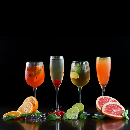 Composition with four different shapes crystal cocktail glasses with cold drinks, orange, lime and grapefruit slices, melting ice cubes, mint leaves and red cherries on black background. Ratio 1:1の写真素材