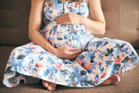 Cropped view of pregnant mother in dress sitting in sofa. Young woman, future mother embracing big tummy and expecting little baby. Concept of birth and happiness.の写真素材