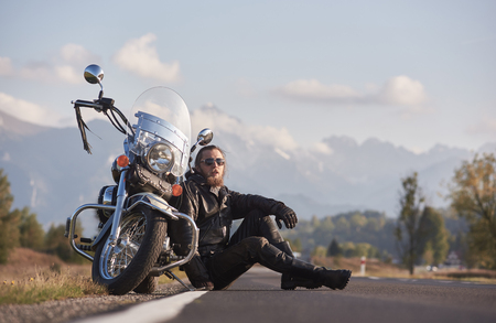 Handsome motorcyclist in black leather clothing sitting at modern powerful shiny motorcycle on grassy roadside on blurred copy space background of distant mountain peaks under bright sky.の写真素材